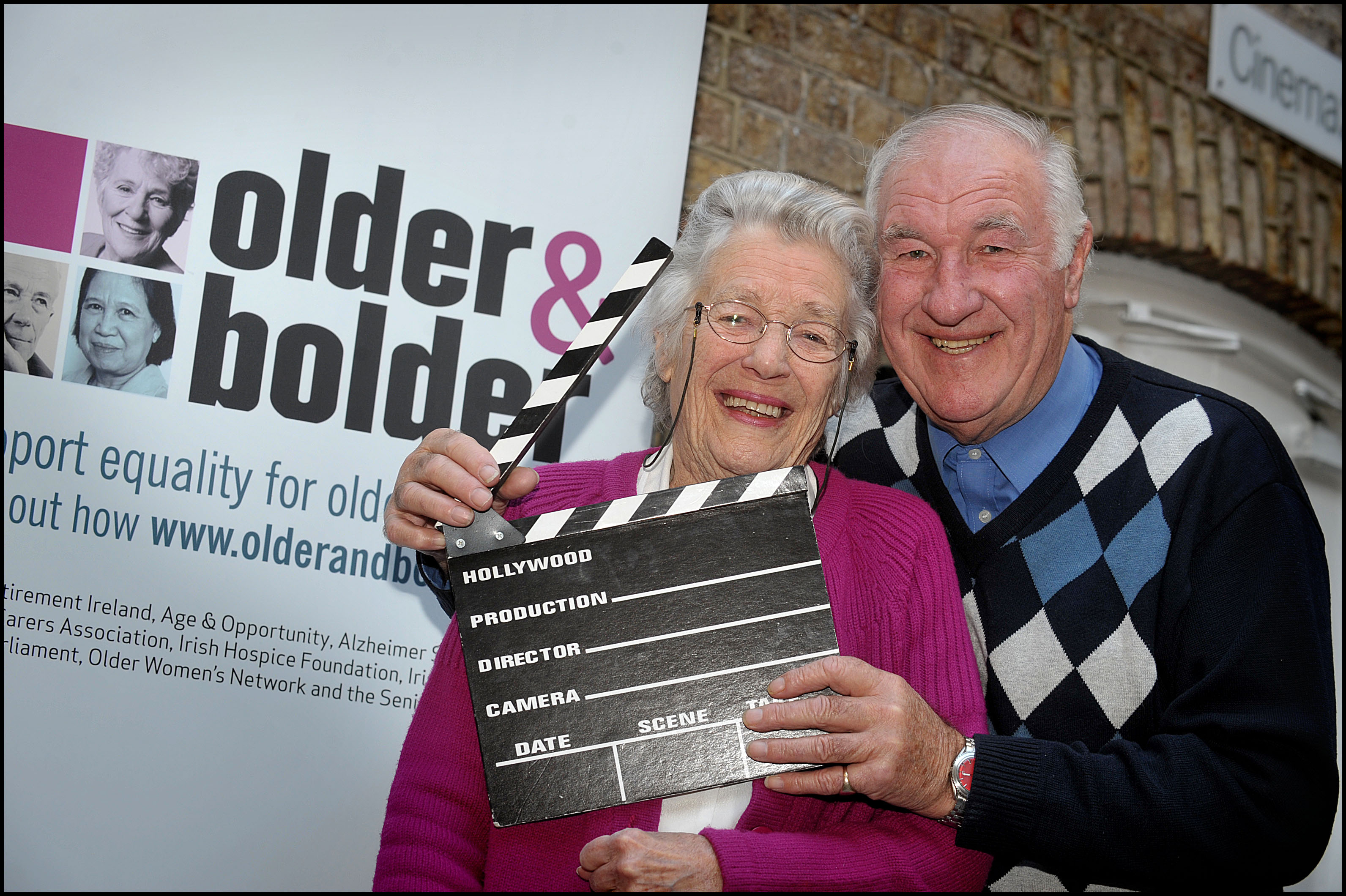 two older people smiling holding a movie clapper-board